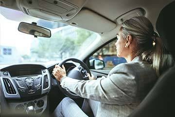 Over the shoulder shot of a woman in a light grey suit driving a car, taken from the back seat