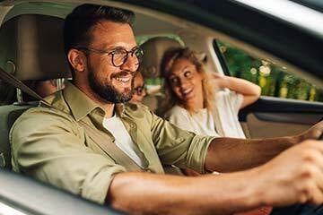 Shot taken from the side of a smiling man driving a car with a woman in the passenger seat