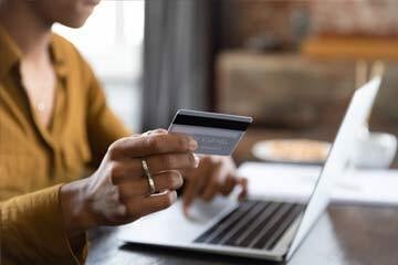 Person typing their bank details into a laptop with one hand while holding their bank card in the other