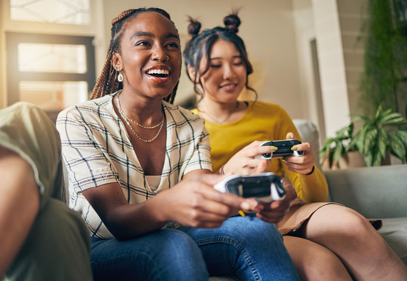 two women sat on a sofa at home playing video games